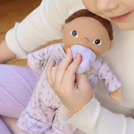 Child holding an Olli Ella Dinkum Doll Binky Baby Pip plush doll with a purple pacifier.