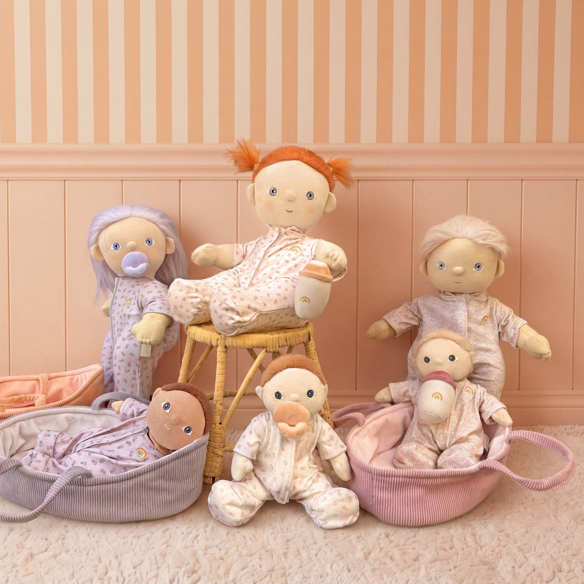 Olli Ella Dinkum Dolls collection in floral pajamas with pacifiers, bottles, and bassinets in a playroom.