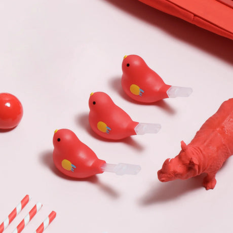 Three red bird-shaped objects with yellow details and plastic spouts, aligned on a pink surface. Nearby are a red ball, striped straws, and a hippo figurine.