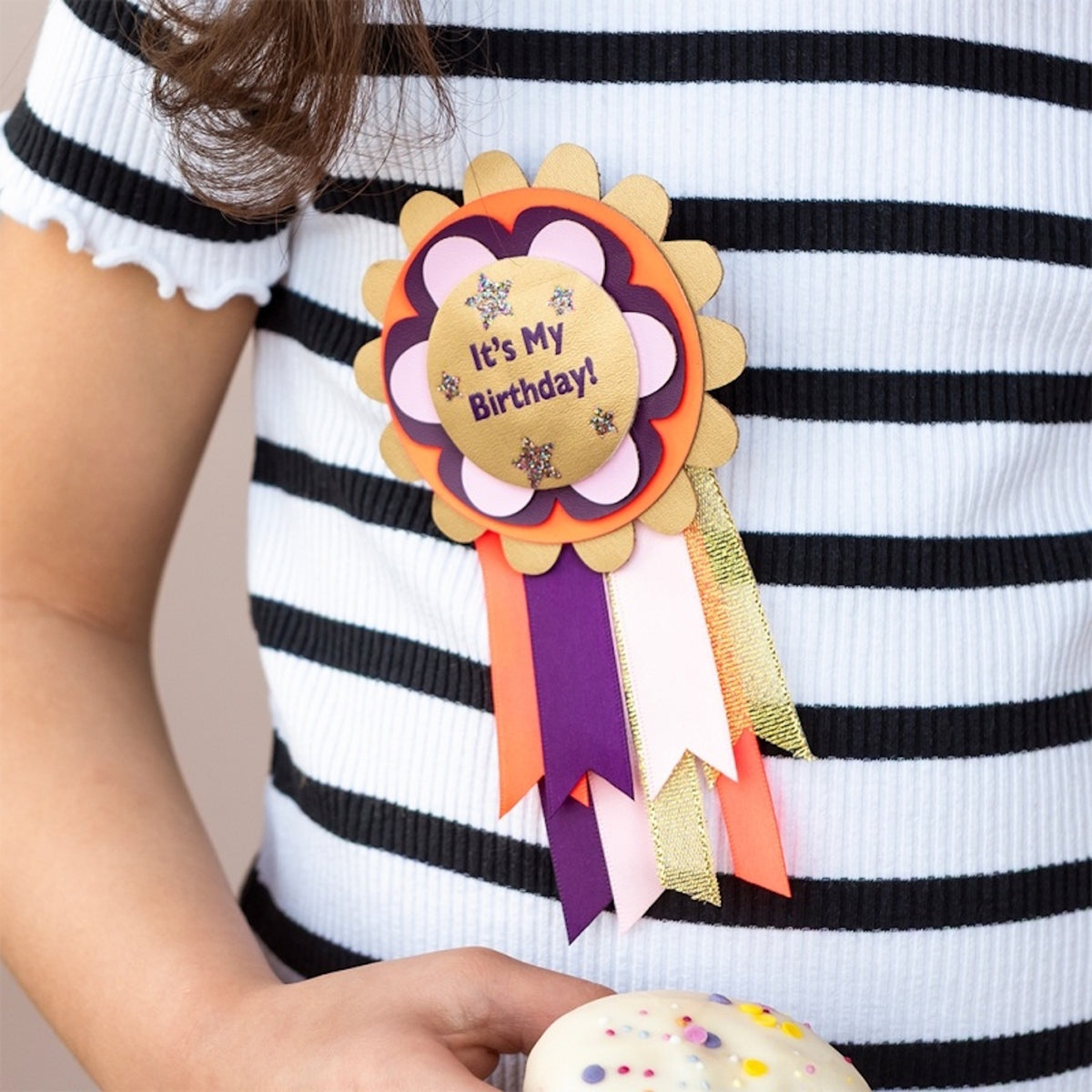 Child in a striped shirt holding a cupcake while wearing an "It's My Birthday!" rosette badge.