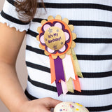 Child in a striped shirt holding a cupcake while wearing an "It's My Birthday!" rosette badge.