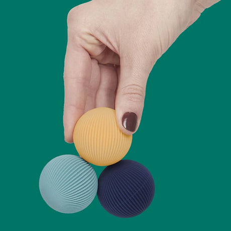 Hand holding three ridged Speks Odds fidget magnets in yellow, teal, and navy against a green backdrop.