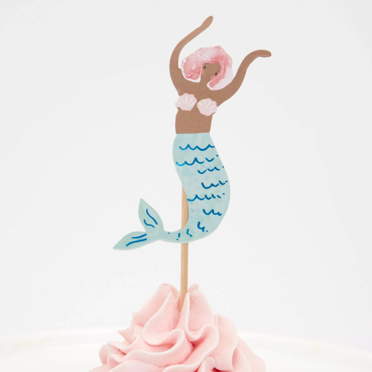 A mermaid cake topper with pink hair and a blue tail, placed on pink frosting against a white background.