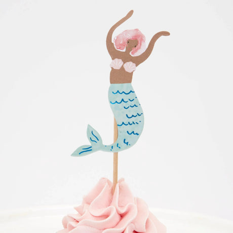 A mermaid cake topper with pink hair and a blue tail, placed on pink frosting against a white background.