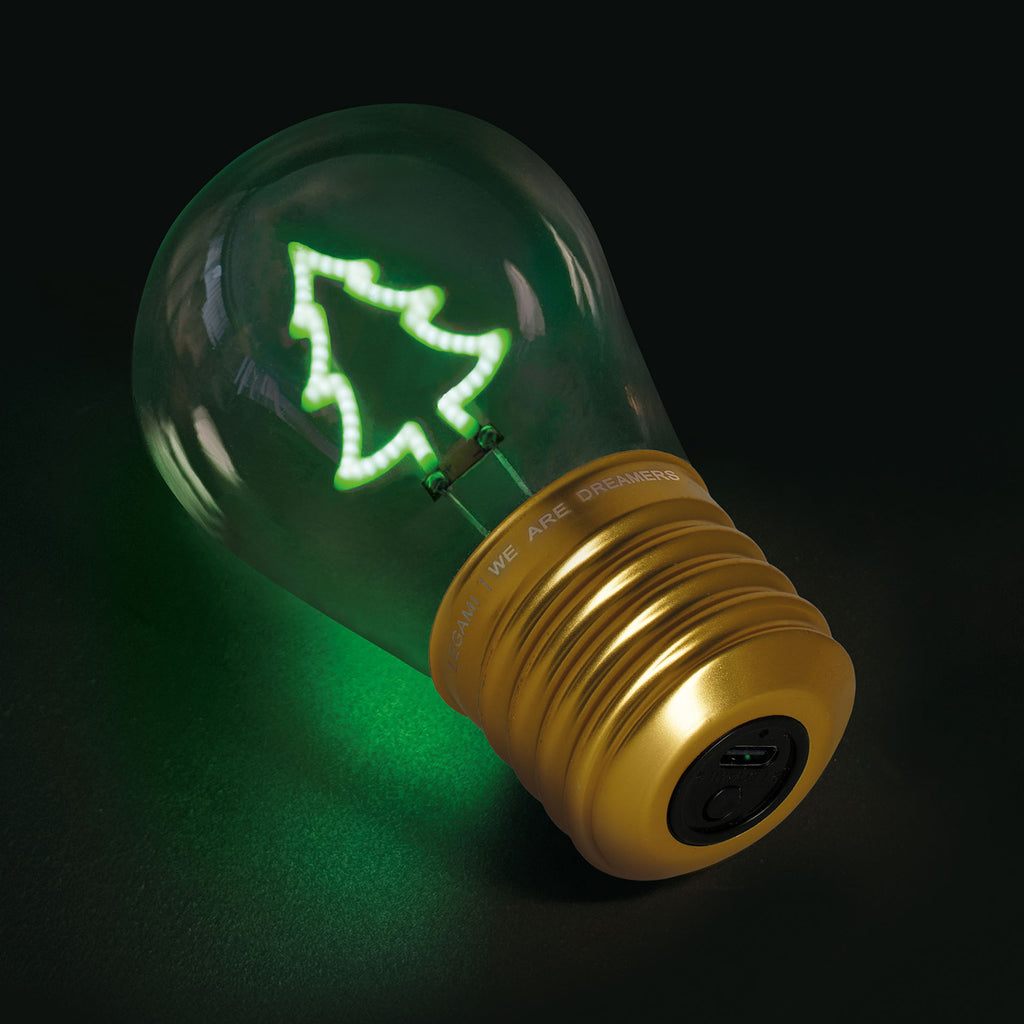 A glowing light bulb with a gold base displays a green LED outline of a Christmas tree inside. The dark background highlights the festive design.