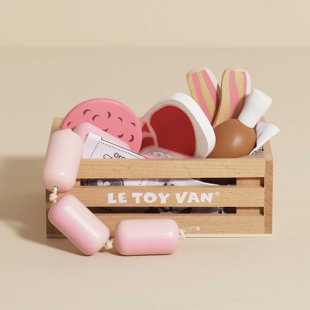 Wooden crate labeled "Le Toy Van" filled with toy food, including pink and white sausages, pizza slices, and drumsticks. Playful and colorful.