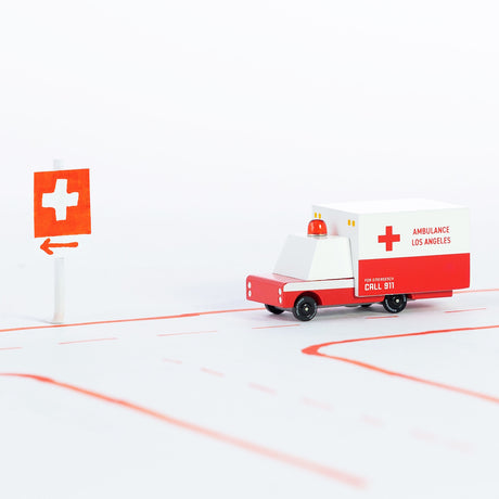 Candylab wooden ambulance toy on a paper road with a red hospital sign, minimalist Los Angeles emergency vehicle design.