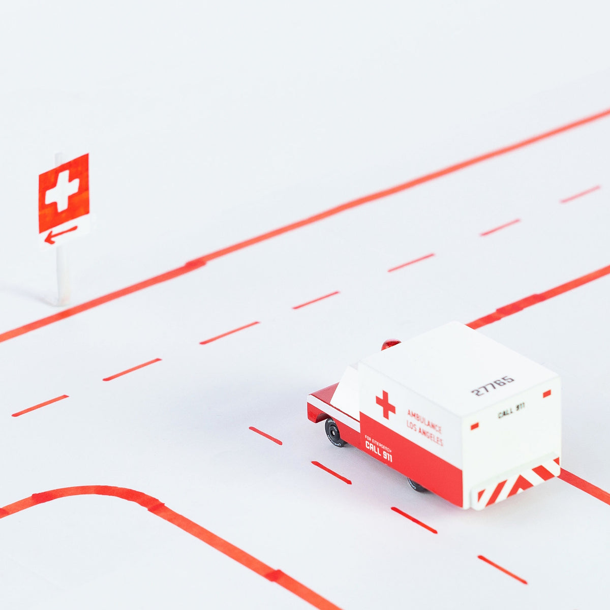A wooden toy ambulance by Candylab Toys driving on a white road with red lines and a medical cross sign.