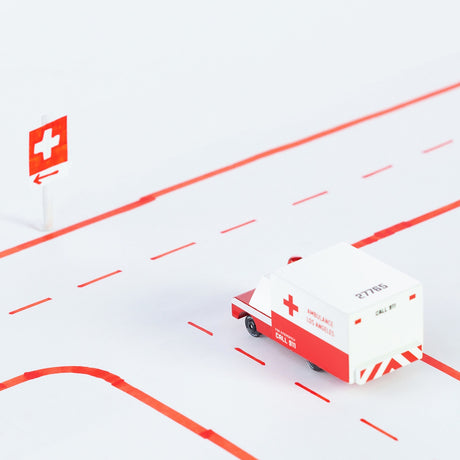 A wooden toy ambulance by Candylab Toys driving on a white road with red lines and a medical cross sign.