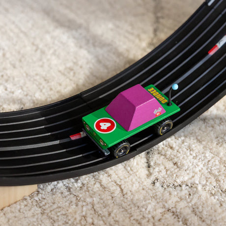 Green and purple Candycar Dundee wooden toy truck racing on a black toy track over a white shag rug.