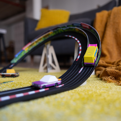 Colorful Candycar wooden toy cars racing on a flexible black track over a yellow rug in a cozy living room.