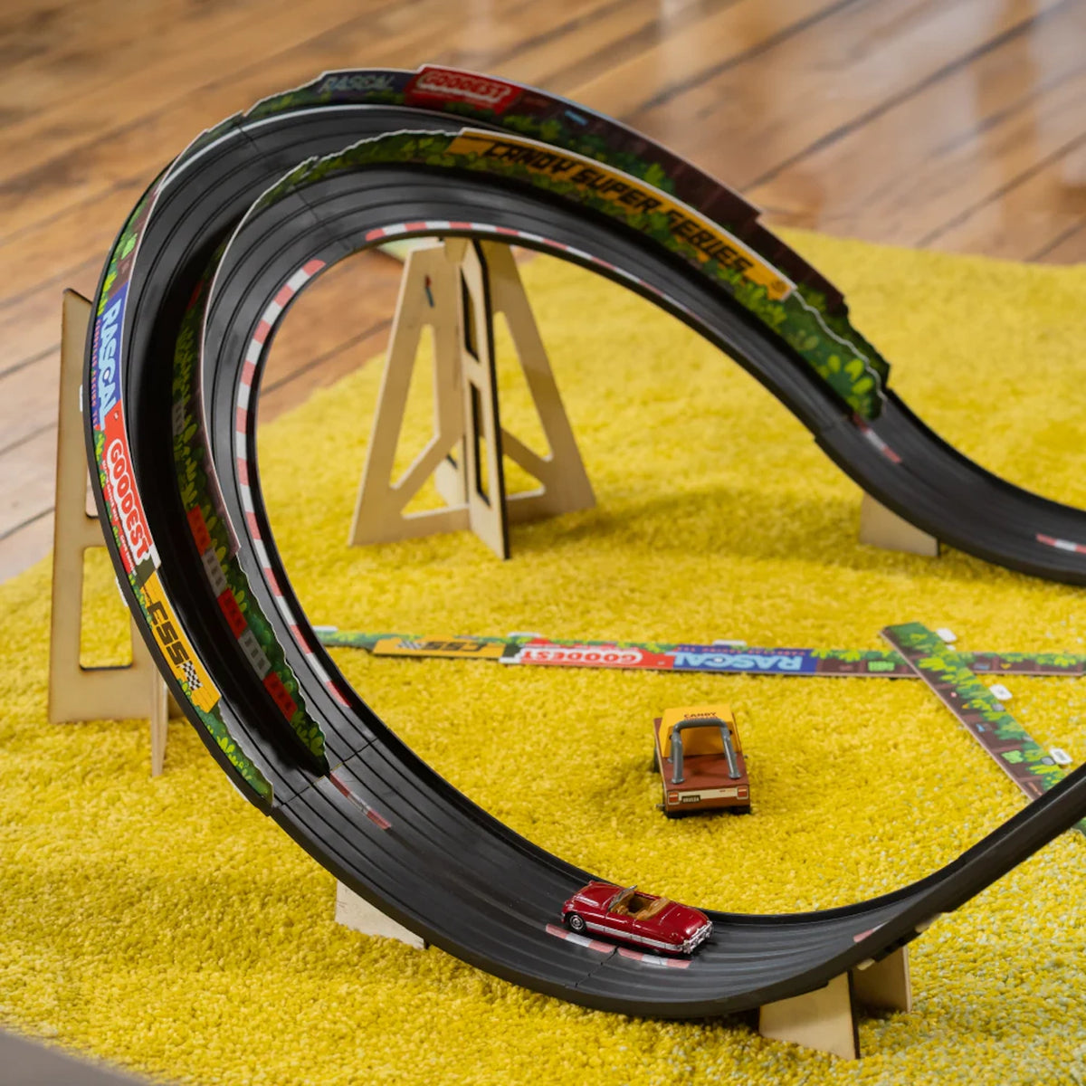 Candylab Roadworks Banks Turn add-on kit with wooden toy cars on a curved black race track over a yellow rug.