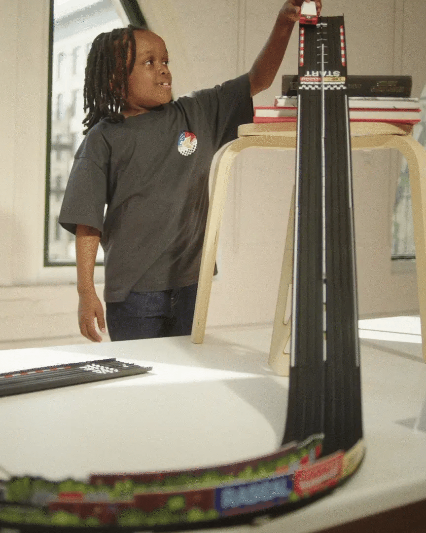 A child plays with a Candylab Roadworks Hairpin Turn kit, racing a toy car down a steep wooden track ramp from a stool.