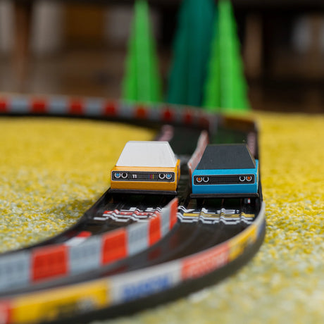 Two toy cars race on a curved wooden track with checkered barriers and green trees on a yellow carpet.