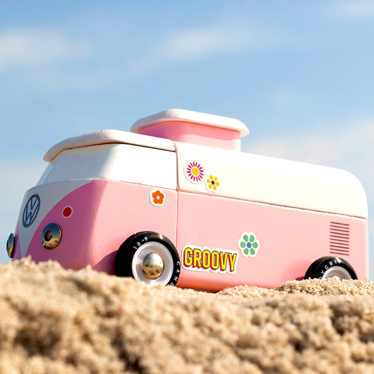Pink toy VW beach van with "Groovy" and flower decals on a sandy beach against a blue sky.