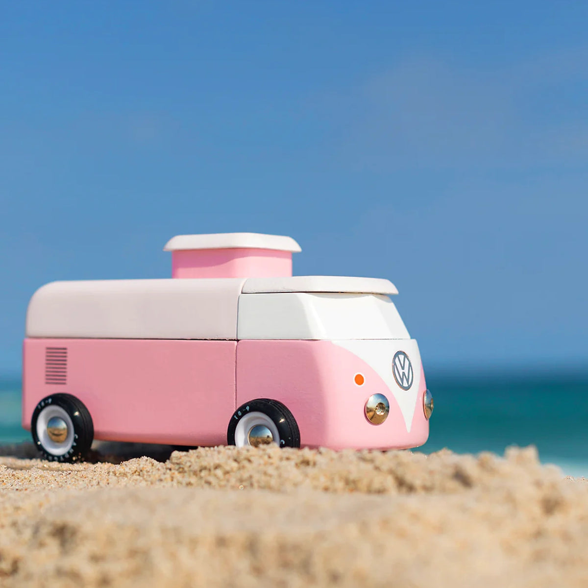 Pink and white wooden toy VW beach bus on a sandy beach with a blue ocean background.