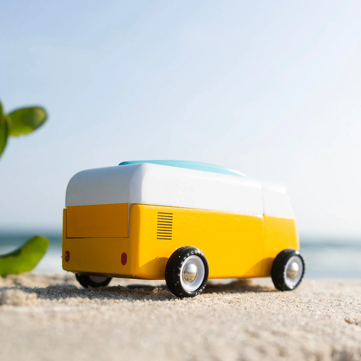 Yellow and white Candylab wooden beach bus toy with a blue surfboard on a sandy beach.