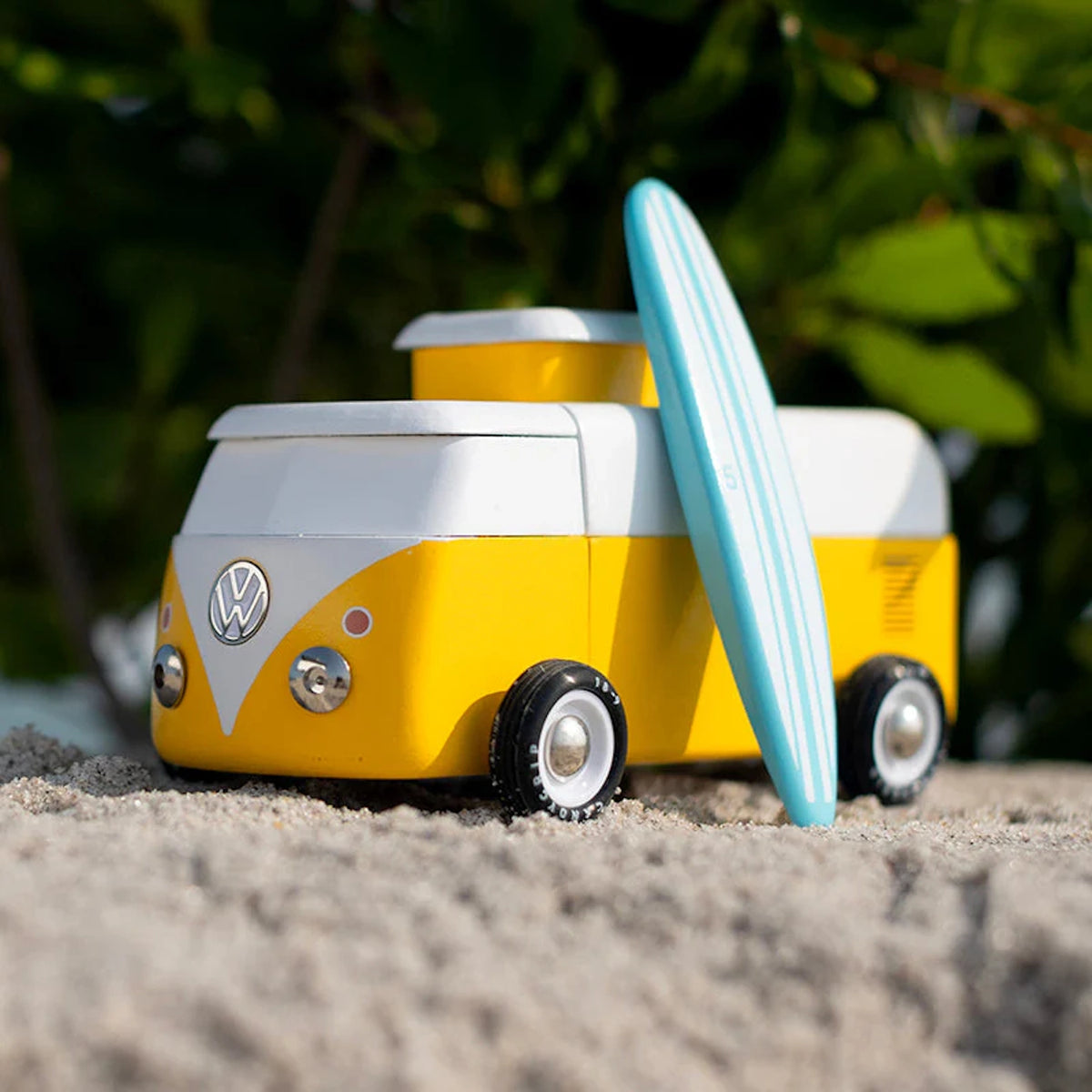 Yellow and white toy Volkswagen beach bus with a blue surfboard on sand.