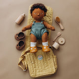 Dinkum doll on a honey yellow changing mat, surrounded by wooden doll accessories, shoes, and a bag.