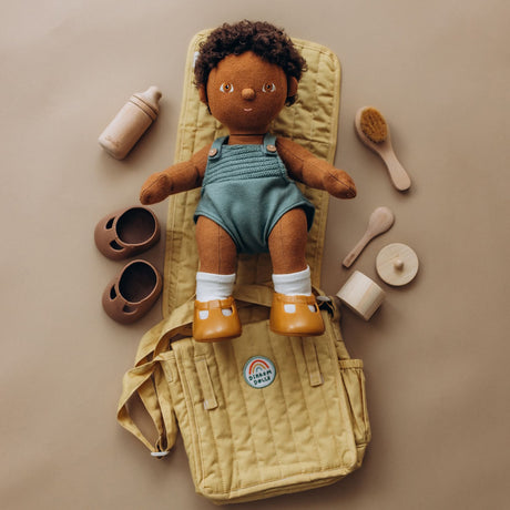 Dinkum doll on a honey yellow changing mat, surrounded by wooden doll accessories, shoes, and a bag.