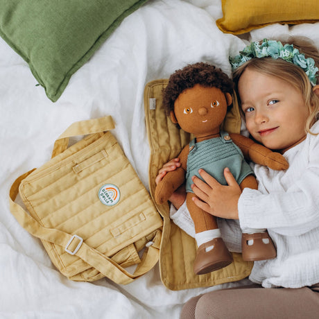 A young girl lying on a bed next to a honey yellow doll changing bag and a Dinkum Doll on a mat.