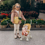 Little girl with a honey-colored crossbody bag standing next to a toy stroller with plush dolls.