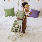 Child holding Dinkum Doll and wearing sage green Carrie convertible changing bag next to doll stroller.