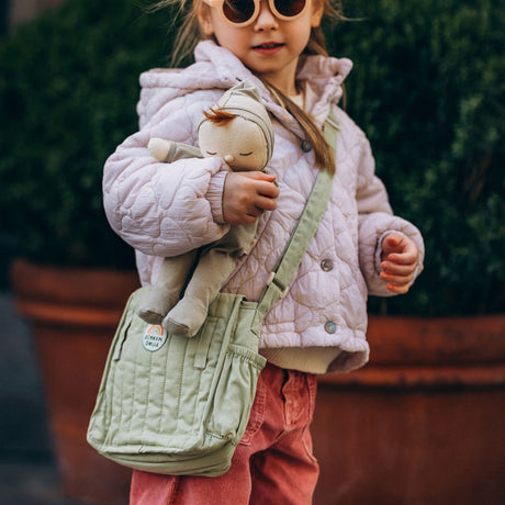 Young girl wearing a sage green quilted diaper bag while holding a Dinkum Doll outdoors.