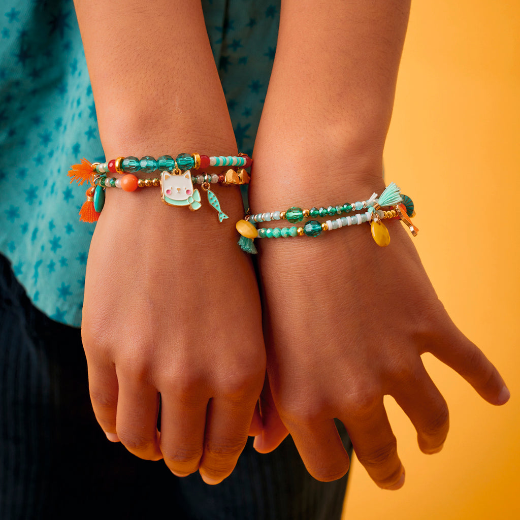 Two hands with colorful beaded bracelets featuring charms, including a cat, against a vibrant orange background. The tone is playful and stylish.