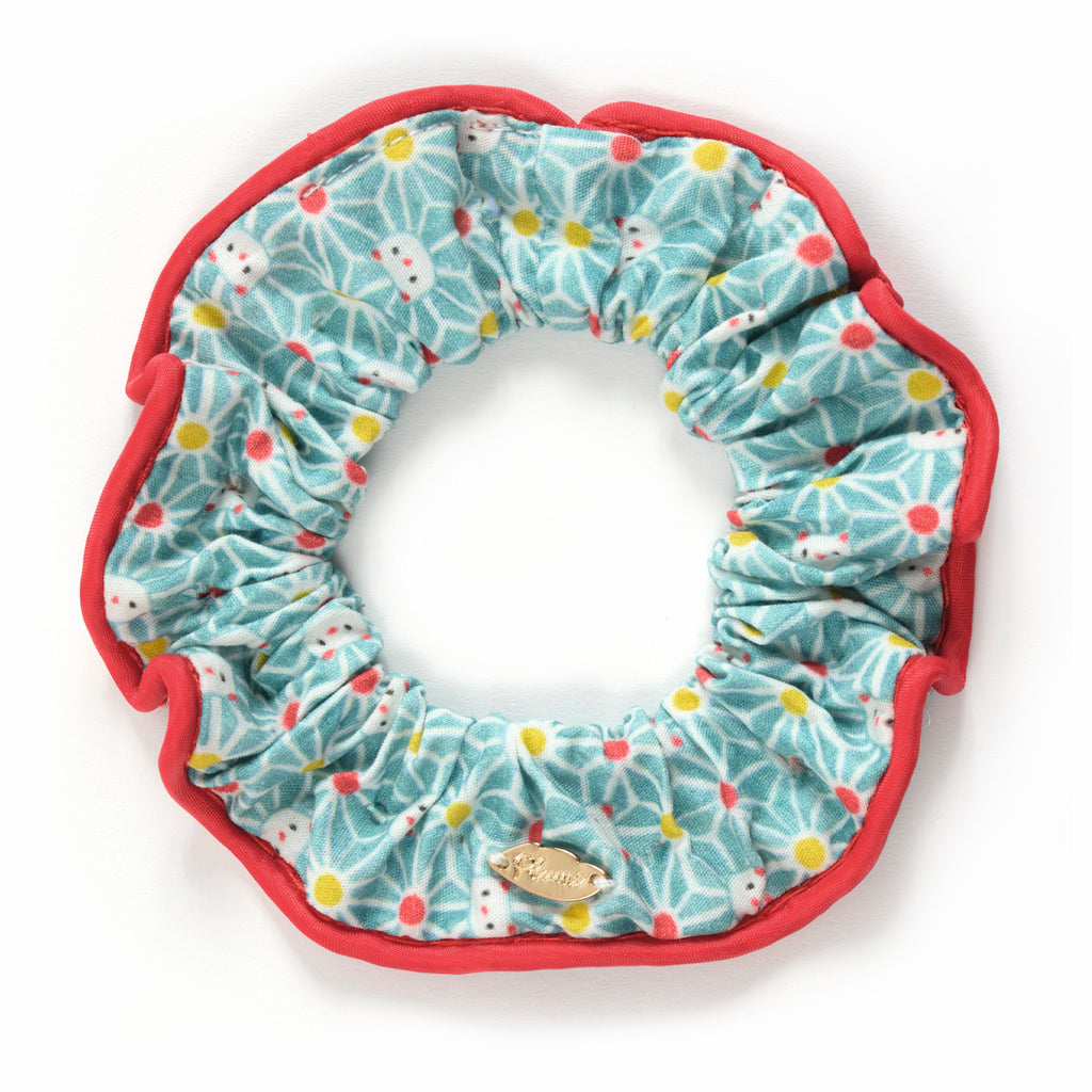 Colorful scrunchie with a floral pattern in blue, red, yellow, and white, accented with a red border. A small gold emblem is attached. Fun and vibrant.