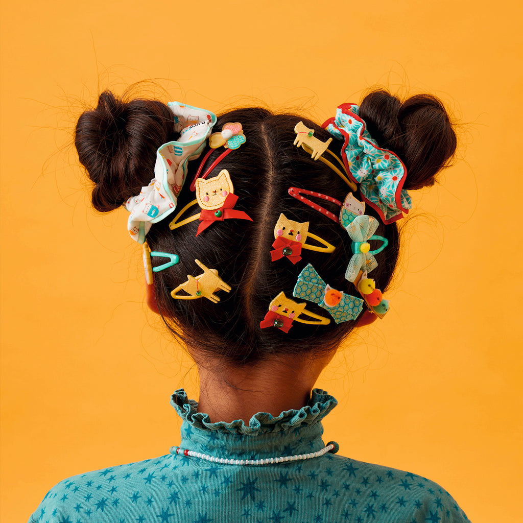 A child's hair styled in two buns with colorful clips and scrunchies featuring playful cat designs against a vibrant orange background.