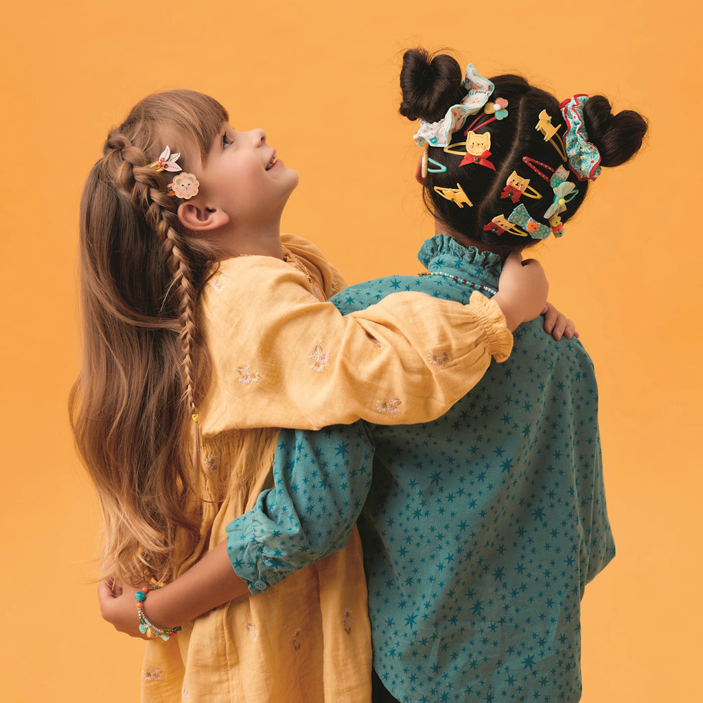 Two young girls stand embraced against a vibrant orange background. One wears a yellow dress with braids, the other in blue with playful hair clips. They convey joy and friendship.