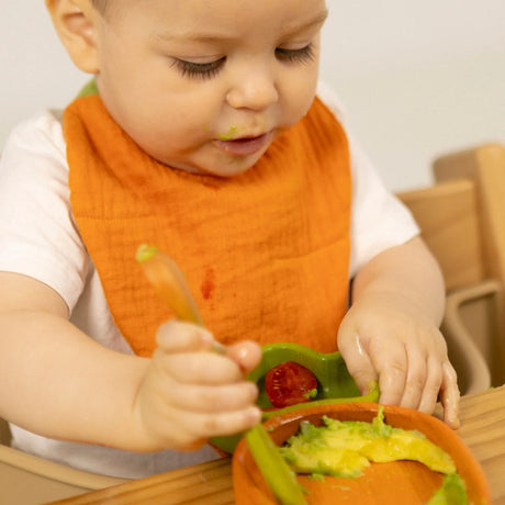Toddler wearing an orange muslin bib while eating avocado and tomato from a wooden bowl in a high chair.