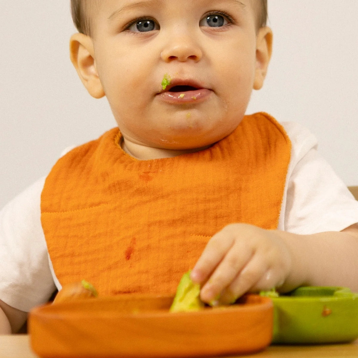 Baby wearing an orange muslin bib while eating avocado from a wooden plate.