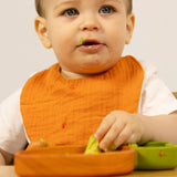 Baby wearing an orange muslin bib while eating avocado from a wooden plate.