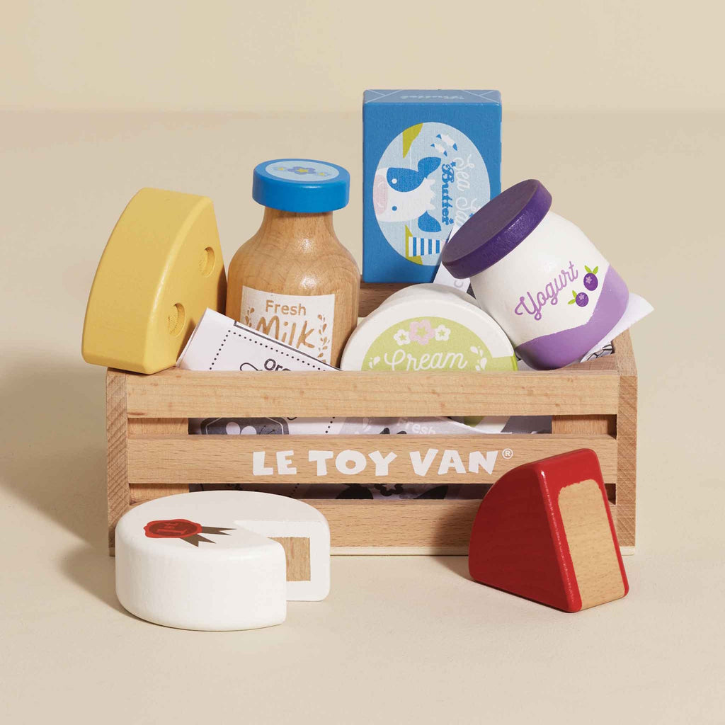 Wooden toy crate labeled "Le Toy Van" containing colorful toy groceries: milk bottle, cheese, yogurt, and cream. Bright, playful, and inviting.