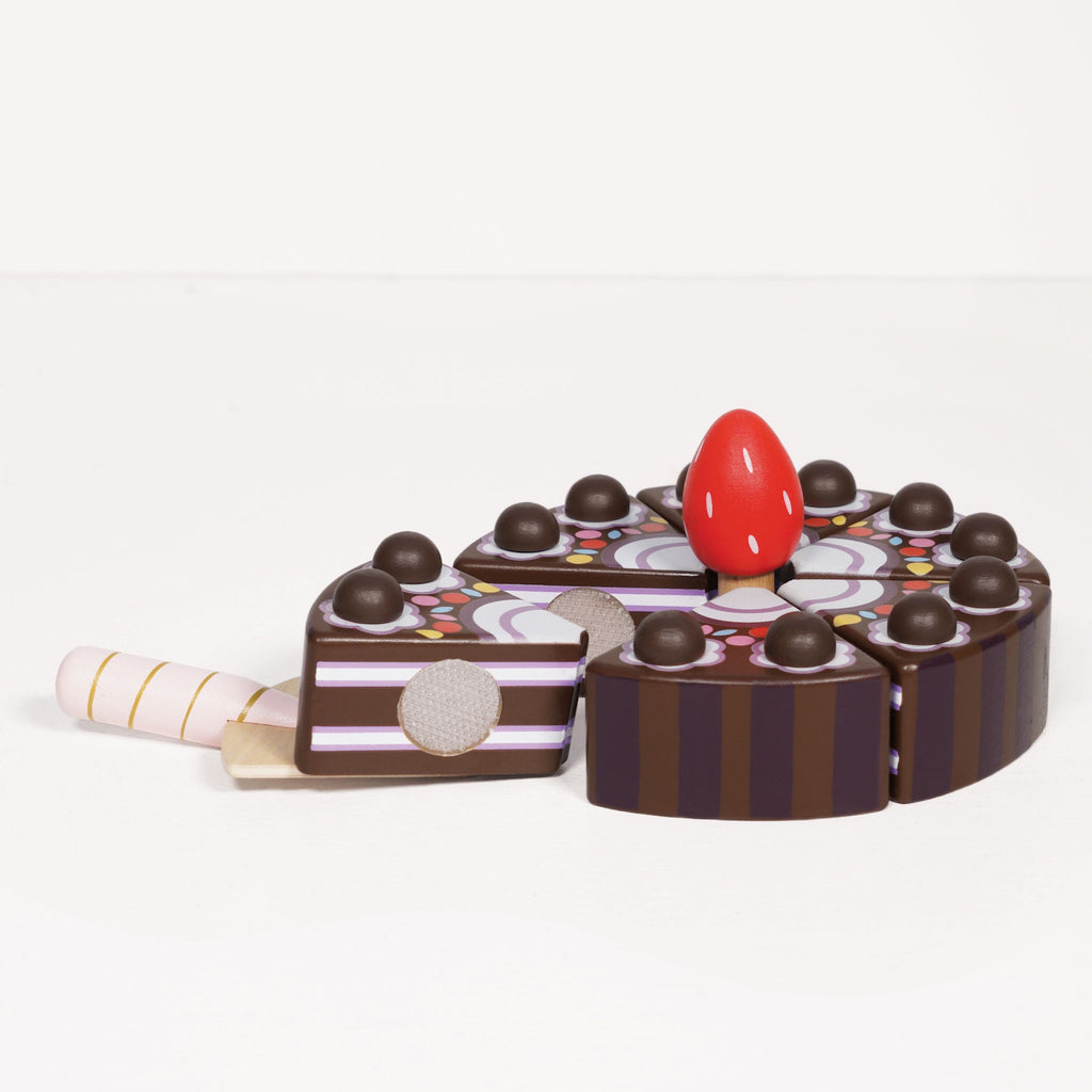 Wooden toy cake sliced into six pieces, decorated with chocolate balls and a red strawberry on top. A pink-striped plastic knife is inserted. Playful and colorful.
