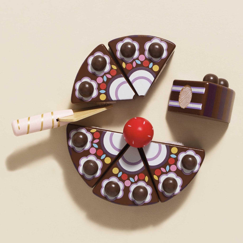 Wooden toy cake sliced into six pieces, decorated with chocolate balls and a red strawberry on top. A pink-striped plastic knife is inserted. Playful and colorful.