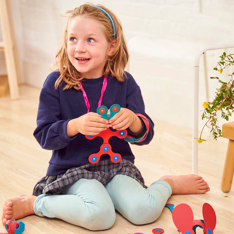 A smiling young girl sits on the floor playing with pink and turquoise magnetic building pieces.