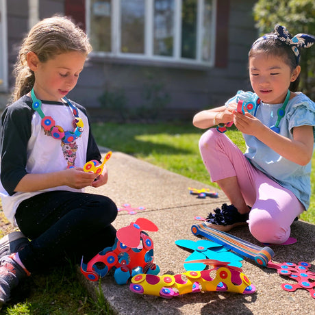 Two young girls sitting outside creating and wearing colorful jewelry made with Clixo Super Rainbow Pack pieces.