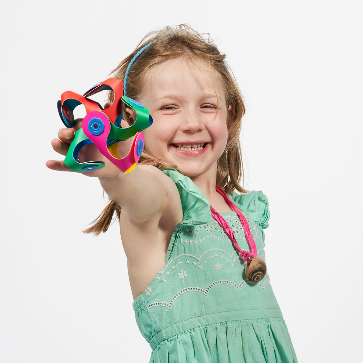 Smiling young girl in a green dress holds out a colorful, rounded Clixo magnetic shape toward the camera.