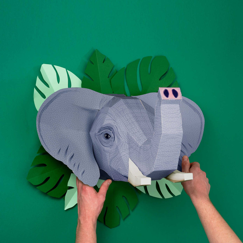 Hands holding a 3D paper elephant head model against a green background with leaf cutouts. The scene conveys creativity and a playful tone.