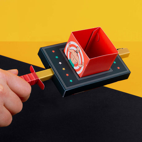 A hand holds a colorful paper craft resembling a magic trick box against a yellow and black background. The red box features a muscleman illustration.
