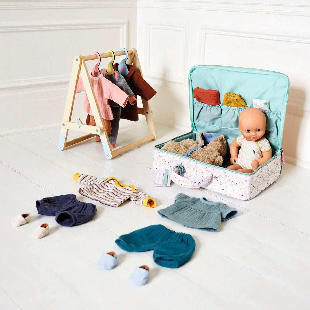 Wooden doll clothing rack and floral travel suitcase with baby doll and miniature outfits on a white floor.