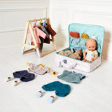 Wooden doll clothing rack and floral travel suitcase with baby doll and miniature outfits on a white floor.