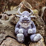 Plush grey horned creature toy sitting outdoors on weathered wooden logs.