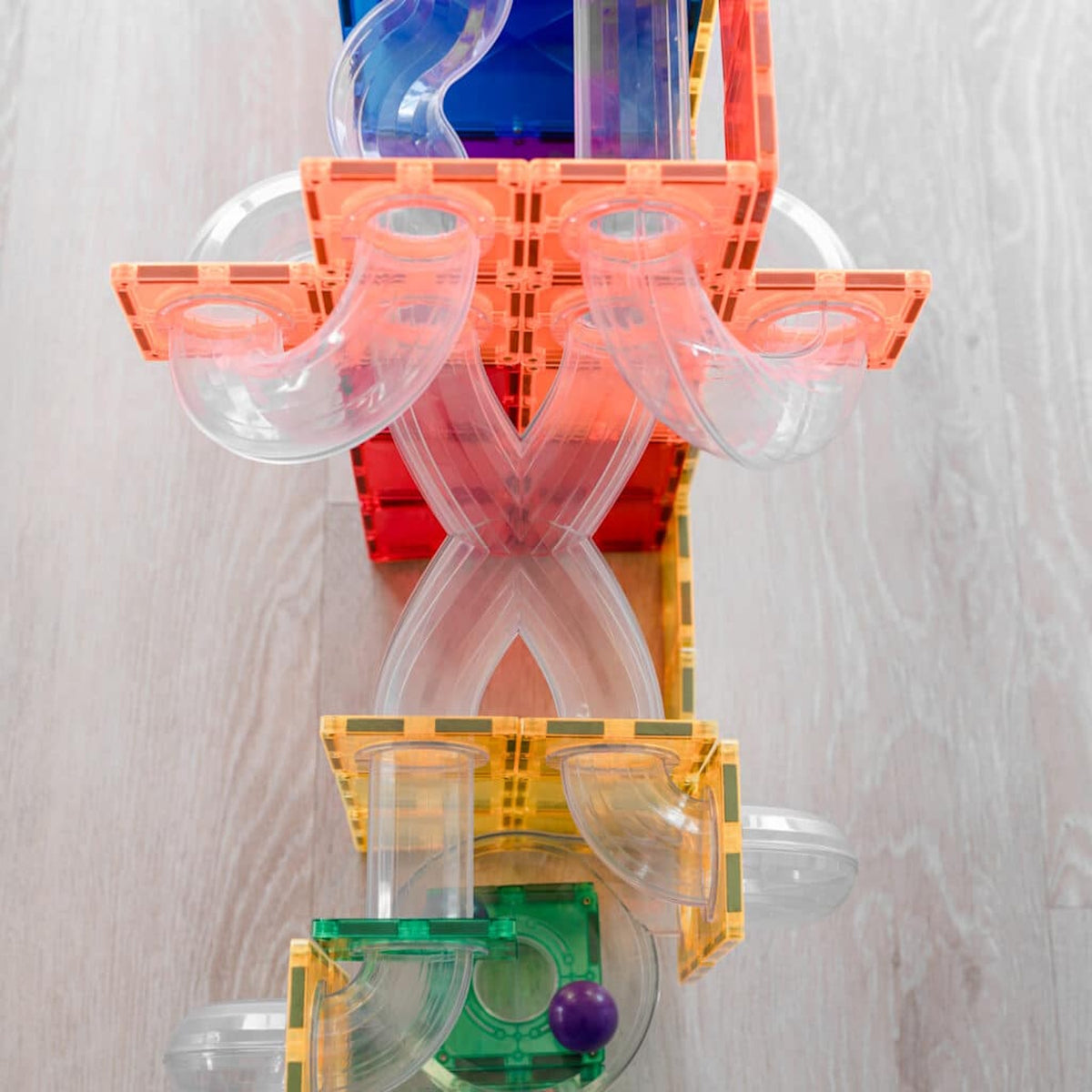 Top-down view of a colorful magnetic tile ball run structure with clear tubes on a light wooden floor.