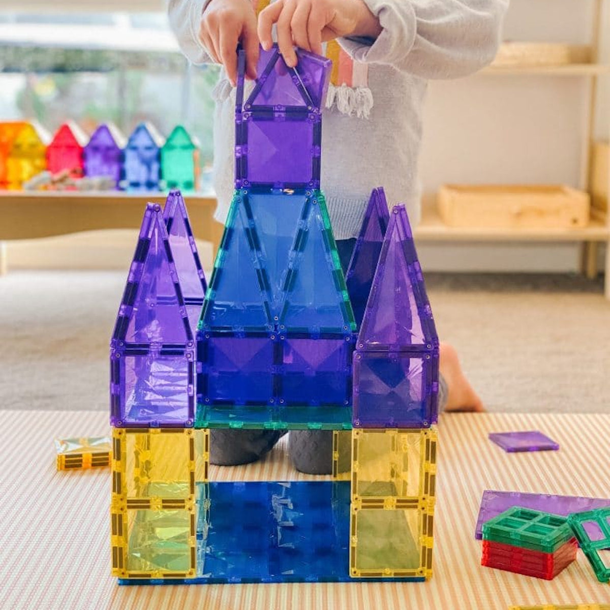 A child builds a colorful toy castle structure using translucent blue, purple, and yellow magnetic tiles.