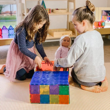 Two young girls playing with dolls on the floor near a structure built with colorful magnetic tiles.