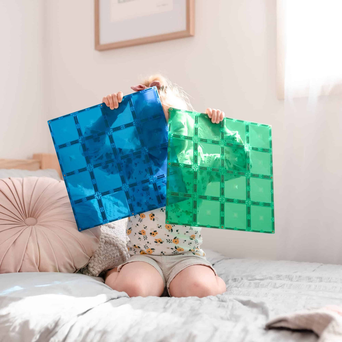A child kneeling on a bed holding large translucent blue and green magnetic tile base plates in front of their face.